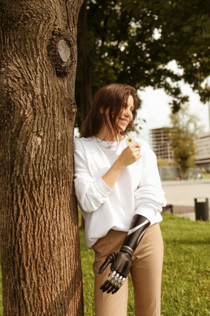 Young woman with prosthetic arm smiling and enjoying nature in a park setting.